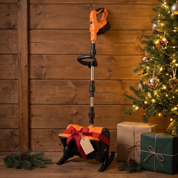 An orange-and-black snow shovel with its shaft folded, wrapped in a loose red ribbon and a plain bow around its head and leaning against a wooden wall beside a lit Christmas tree, a blank kraft tag on the ribbon and wrapped boxes on the floor